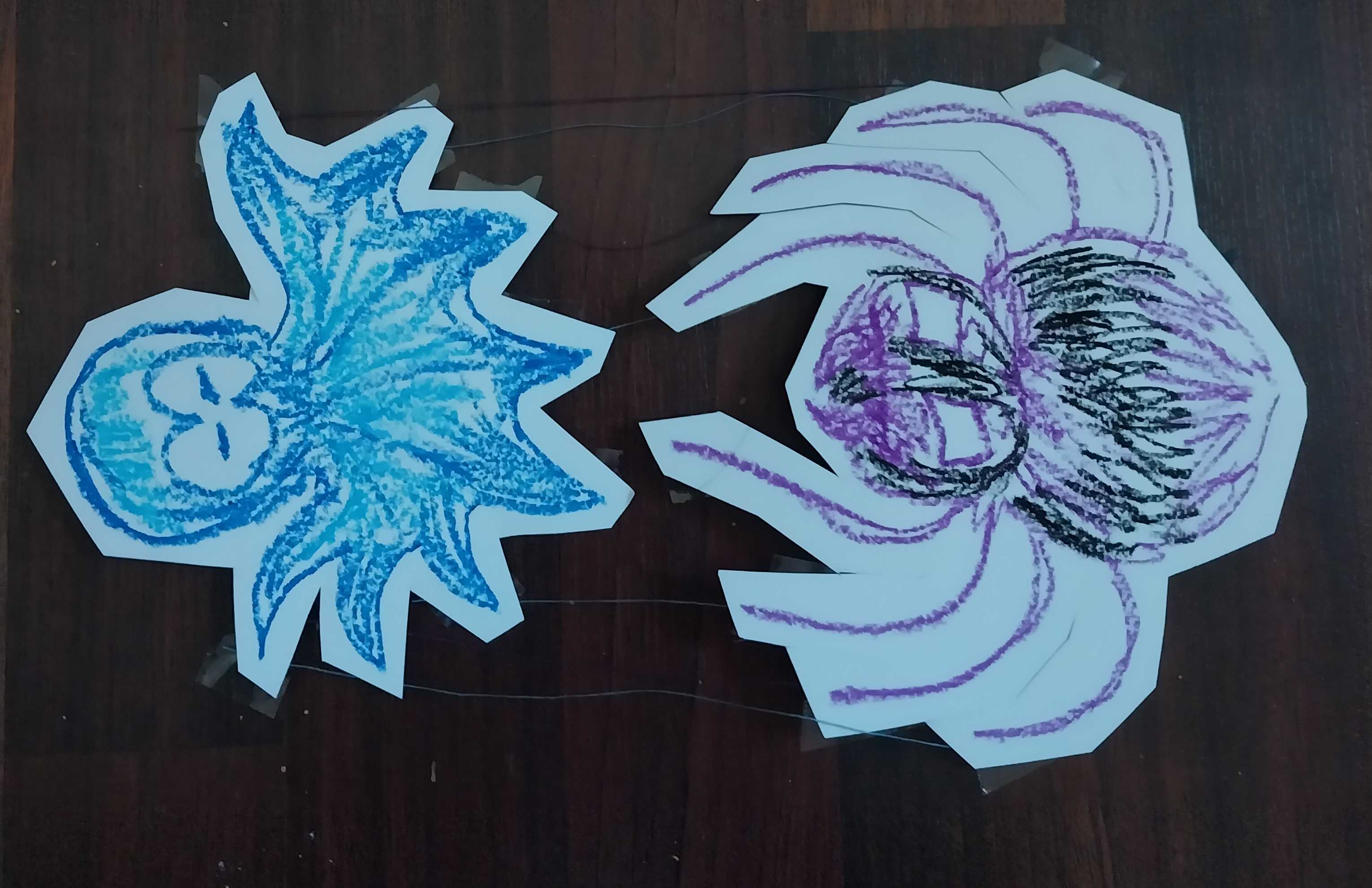 A photo of two cardstock papercrafts. One craft is an octopus colored blue with pastel and the other is a spider colored black and purple. Their legs are connected by string.